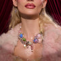 Glossy-lipped model showcases glitter-dusted chest and a chunky pastel beaded necklace with pearls and charms, wearing star earrings and a pink faux-fur coat against red velvet drapes, delivering a ColourPop holiday glam look.