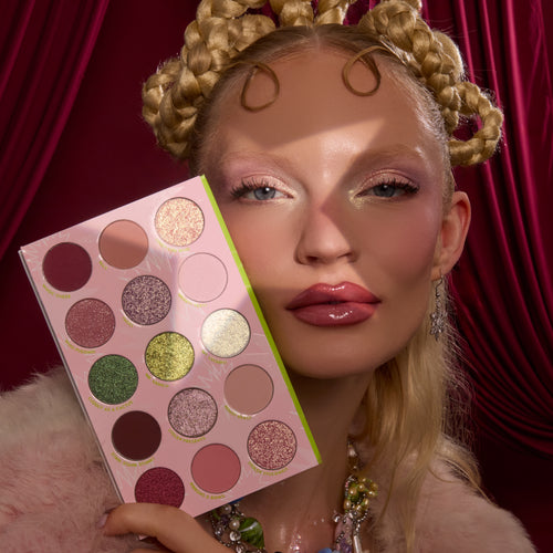 How the Grinch Stole Christmas Shadow Palette by ColourPop held beside a person’s glam face, showing a pink 16-pan mix of matte and glitter eyeshadows - greens, pinks, golds, and plums - set against rich red drapery for a festive holiday look.