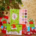 ColourPop x The Grinch eyeshadow palette opens to 15 holiday shades - pinks, greens, and golds in matte and shimmer - displayed with Grinch artwork, set among Christmas ornaments and wrapped gifts for a festive makeup scene.