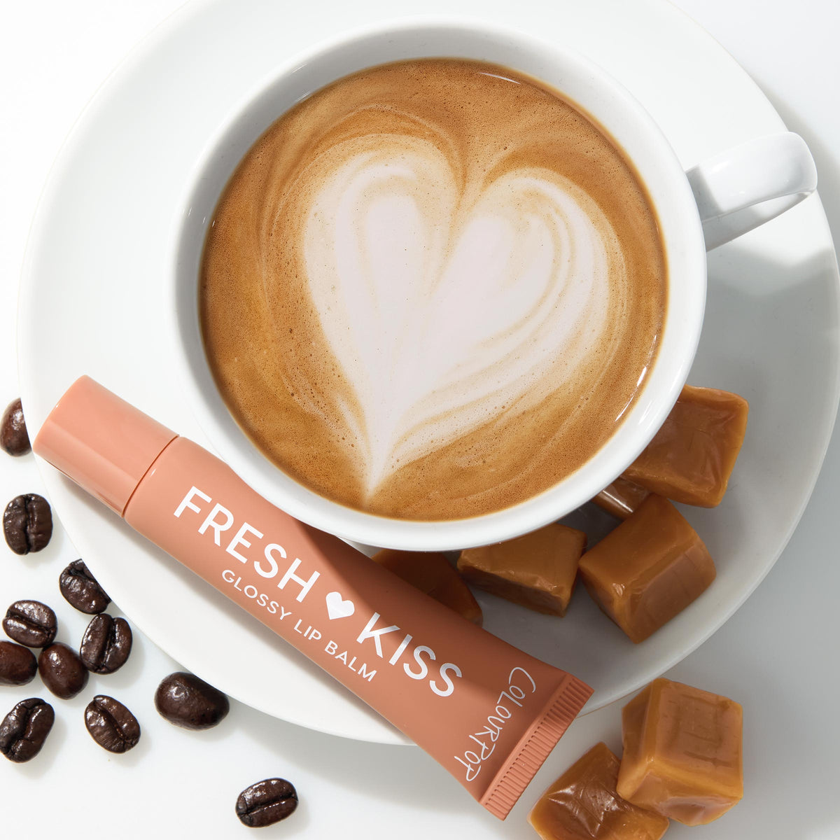 ColourPop Fresh Kiss Glossy Balm in Caramel Latte lies across a white saucer beside a latte with heart foam art, styled top-down with caramel candies and coffee beans, highlighting the nude-caramel tube and glossy lip balm packaging.