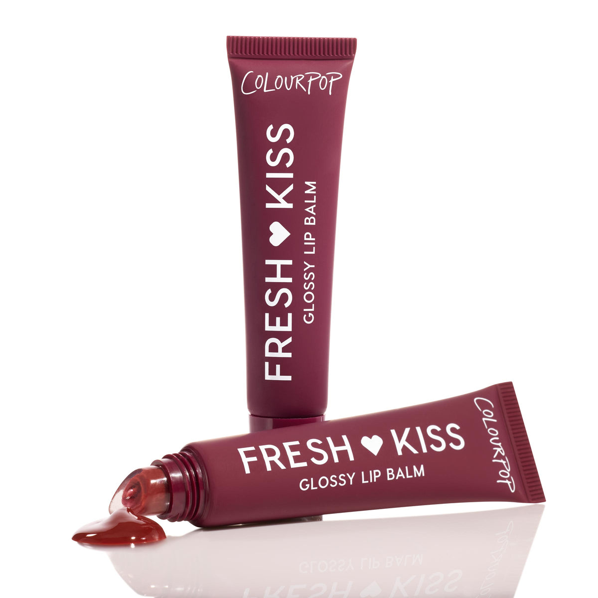 ColourPop Fresh Kiss Glossy Balm in Wild Berry stands upright while a second maroon squeeze tube lies uncapped with glossy berry tint pooling at the tip, on a white reflective studio background.