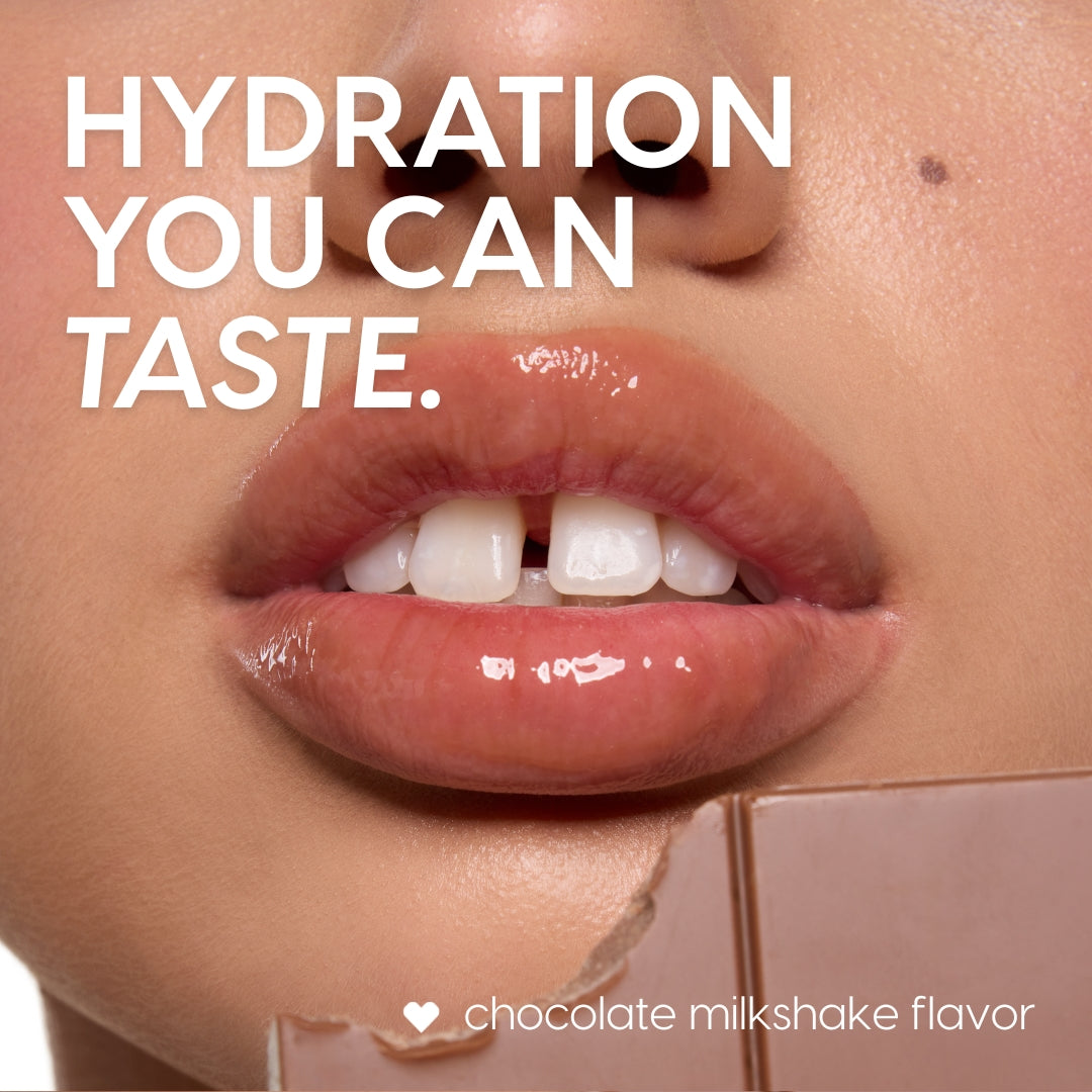 Glossy, smooth lips showcase the ColourPop Lip Scrub - Chocolate Milkshake, with a chocolate bar held near the mouth in a tight beauty close-up, emphasizing the chocolate milkshake flavor and a soft, freshly exfoliated finish.