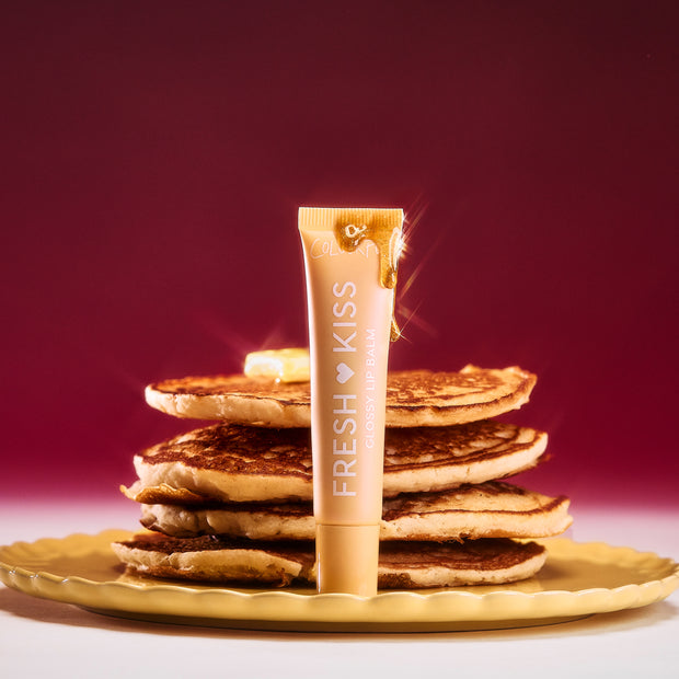 Colourpop Fresh Kiss Glossy Balm in Buttery Pancakes flavor standing upright against a stack of syrup-drizzled pancakes on a yellow plate, styled on a rich burgundy background to showcase this Lip Gloss & Lip Oil hybrid.