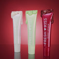 Three Colourpop Holiday Drip Fresh Kiss Glossy Balm tubes stand upright on a reflective surface, pearl, mint, and ruby red with glittery balm dripping from the caps, styled against a glowing red background to showcase festive value sets.