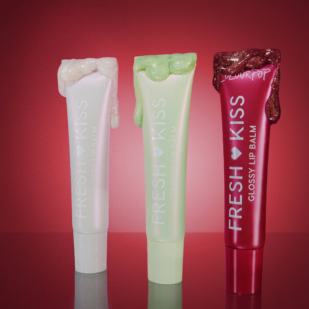 Three Colourpop Holiday Drip Fresh Kiss Glossy Balm tubes stand upright on a reflective surface, pearl, mint, and ruby red with glittery balm dripping from the caps, styled against a glowing red background to showcase festive value sets.
