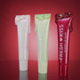 Three Colourpop Holiday Drip Fresh Kiss Glossy Balm tubes stand upright on a reflective surface, pearl, mint, and ruby red with glittery balm dripping from the caps, styled against a glowing red background to showcase festive value sets.