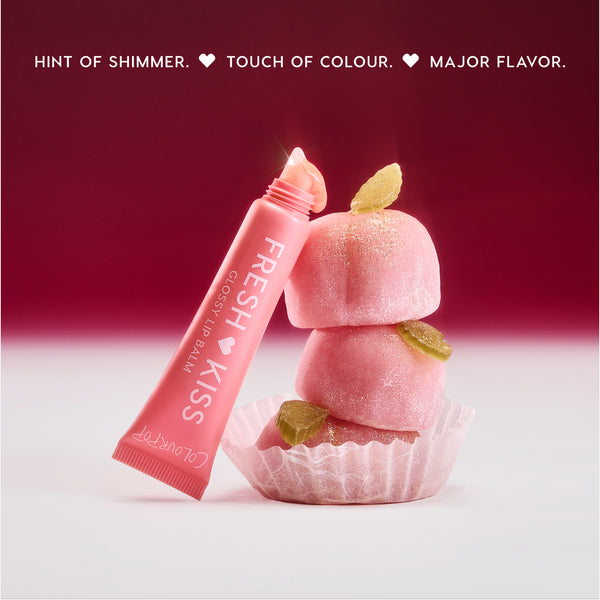 Colourpop Fresh Kiss Glossy Balm in Peach Mochi leaning against stacked pink mochi candies with sugar crystals, highlighting the shimmery Lip Gloss & Lip Oil hybrid on a soft red and white background.