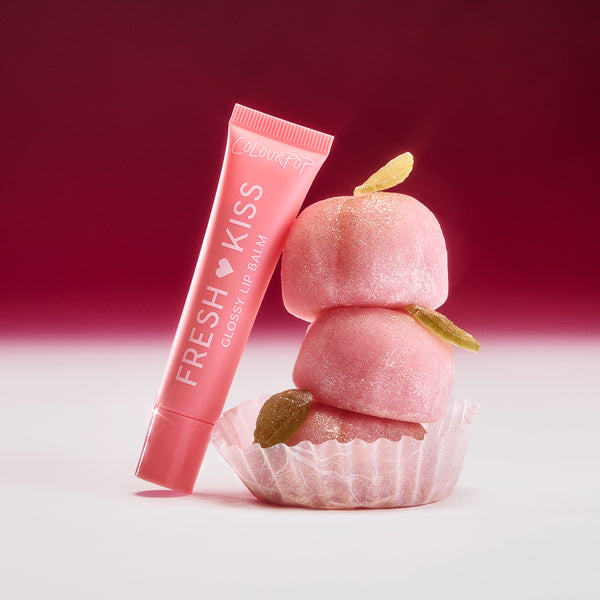 Colourpop Peach Mochi Fresh Kiss Glossy Balm tube leans against a stack of sugar-dusted pink mochi candies on a white surface with rich red gradient background, highlighting the lip gloss & lip oil hybrid’s sweet, dessert-inspired shade.