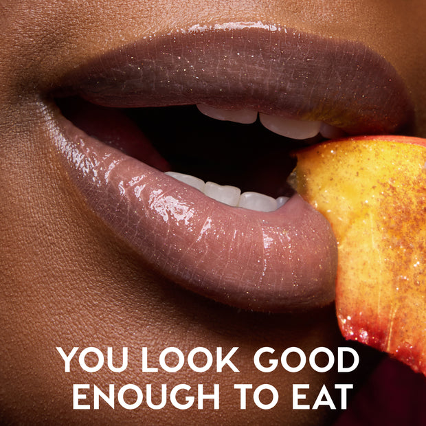 Close-up of shiny, peachy nude lips wearing Colourpop Fresh Kiss Glossy Balm in Peach Mochi, lightly parted as they bite into a juicy peach slice, promoting Lip Gloss & Lip Oil with the tagline “You look good enough to eat.”