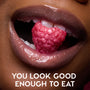 Glossy, shimmer-coated lips wearing Colourpop Fresh Kiss Glossy Balm in Raspberry Crème hold a sparkling raspberry between white teeth, promoting sweet, juicy Lip Gloss & Lip Oil shine with the caption “You look good enough to eat.”