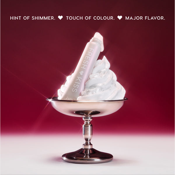 ColourPop Fresh Kiss Glossy Balm in Whipped Cream shade resting in a swirl of shimmering whipped cream in a silver dessert cup against a red gradient background, highlighting the glossy balm as a hybrid Lip Gloss & Lip Oil product.