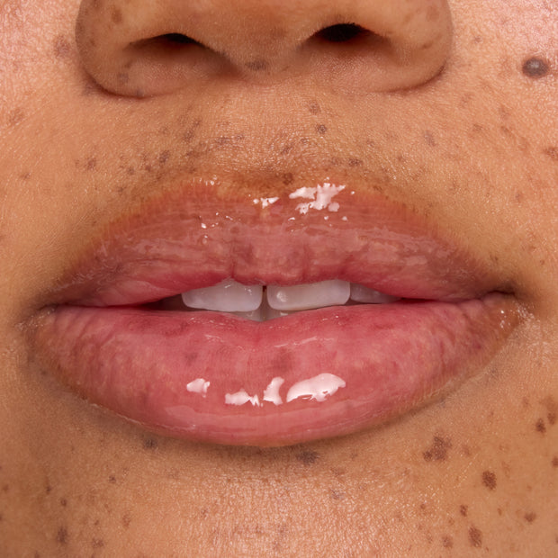 Glossy, hydrated lips wear a clear lip mask with small white balm flecks melting on the surface, shown in an extreme close-up on freckled skin to showcase ColourPop lip mask treatment shine and texture.