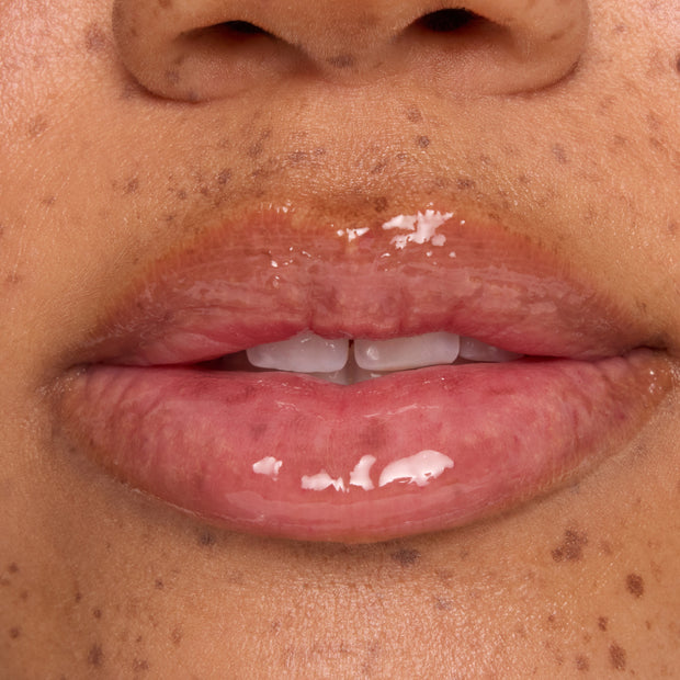 Glossy pink lips wearing a clear, high-shine lip treatment, captured in a tight macro on freckled skin; hydrating sheen and smooth texture showcased as a ColourPop beauty swatch for a plump, moisturized finish.
