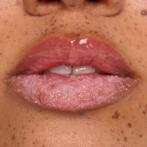 Glossy pink lips exfoliating with a sugar‑crystal lip scrub, bottom lip coated in grainy treatment while the top lip shines with balm, showcasing a ColourPop lip care product in a beauty demo against natural freckled skin.