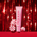 ColourPop Fresh Kiss Glossy Lip Balm in a pink squeeze tube stands upright beside red-and-white peppermint candies on a red surface, framed by sparkling red tinsel for a festive holiday beauty gift scene.