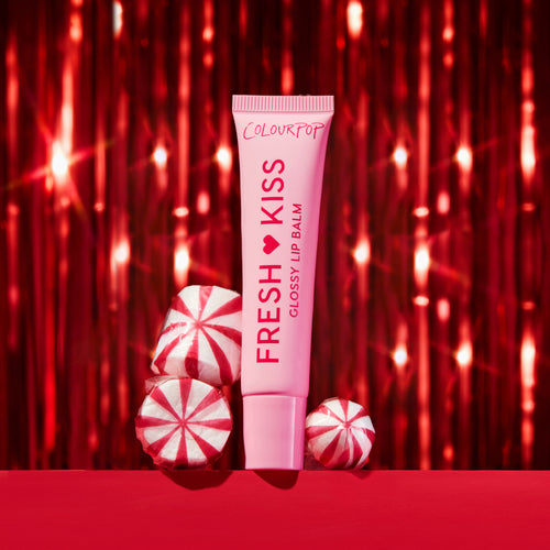 ColourPop Fresh Kiss Glossy Lip Balm in a pink squeeze tube stands upright beside red-and-white peppermint candies on a red surface, framed by sparkling red tinsel for a festive holiday beauty gift scene.