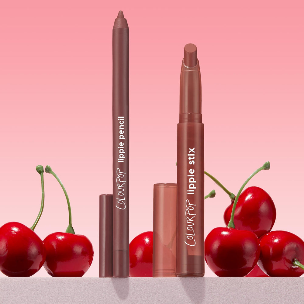 ColourPop Lippie Stix + Pencil Set Big Smooch stands uncapped, revealing a rosy brown lipstick and matching liner with caps nearby, styled among glossy cherries against a soft pink gradient backdrop.