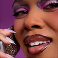 Model smiles while talking on a flip phone, wearing Heart to Get 2 Die 4 ultra glossy lip in a deep brown shine, paired with bold pink and purple heart-shaped eye makeup against a vibrant purple background.