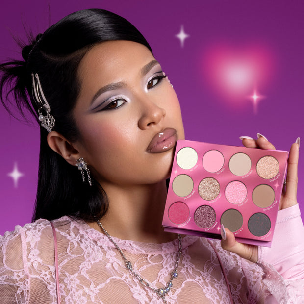 Model with bold pastel eye makeup holds the Heart to Get Full Collection eyeshadow palette, showing 12 shimmery and matte pink, nude, and berry shades against a glowing purple background, promoting the Heart to Get full collection set.