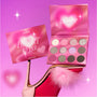 ColourPop Heart to Get Full Collection eyeshadow palette opened to show 12 shimmery and matte pink, nude, and glitter shades, styled on a pink high heel with feathers against a glowing pink background for the Heart to Get makeup set.