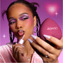 Model applies shimmery pink gloss while holding a heart-shaped ColourPop compact, showing bold purple and pink heart eye makeup that highlights the Heart to Get Full Collection full collection set against a dreamy pink background.