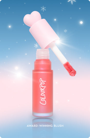 Colourpop liquid blush with a heart-shaped pink cap, doe-foot applicator lifted from a frosted coral bottle showing creamy pigment, set against a blue-to-pink snowy holiday backdrop for a festive, gift-ready look.