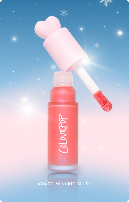 Colourpop liquid blush with a pink heart-shaped cap and doe-foot applicator lifting coral pigment from a frosted bottle, set against a blue-to-pink snowy holiday backdrop, highlighting award-winning makeup for US shoppers.