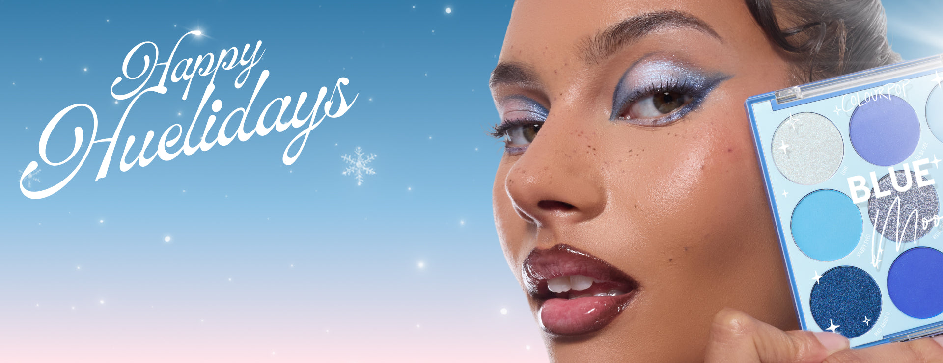Colourpop Blue Moon nine-pan eyeshadow palette held beside a model's face, showing icy blue shimmer eyeshadow and glossy lips, against a snowy gradient sky with 'Happy Huelidays' script - festive holiday makeup ad in cool-toned blues.