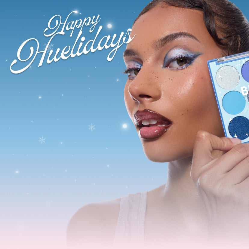 Colourpop blue eyeshadow palette held to a model’s cheek, showing icy metallic and matte blues, as she wears bold frosty blue winged shadow and glossy lips against a snowy gradient backdrop with “Happy Huelidays” script.