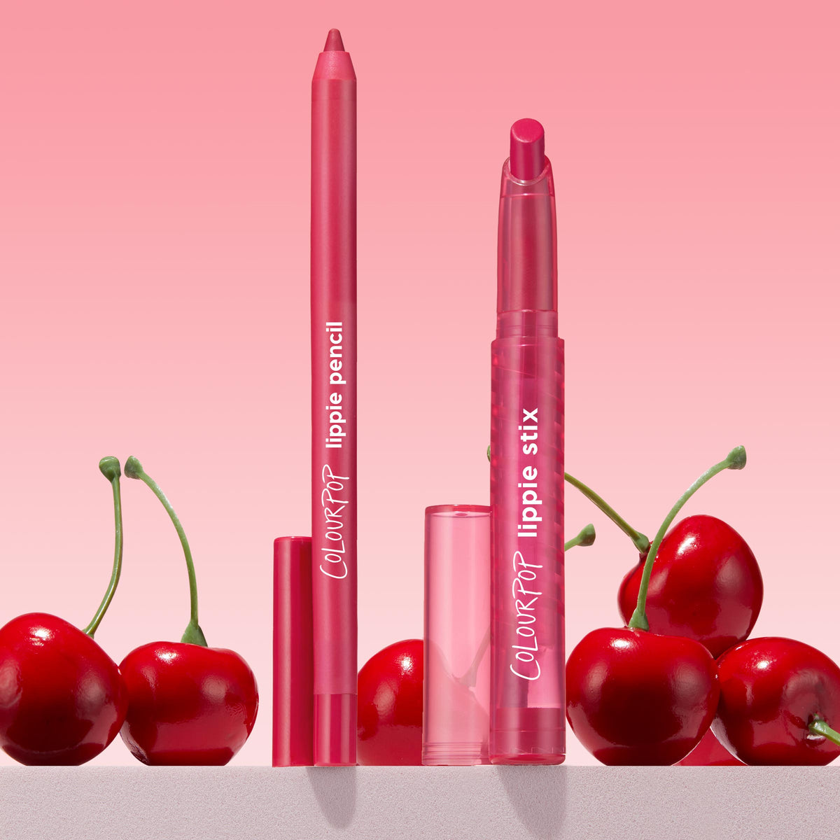 ColourPop Lippie Pencil in I Heart This stands upright with tip exposed beside a matching Lippie Stix, both in vivid cherry-pink, clear caps set nearby, styled with glossy cherries on a light surface against a pink gradient background.