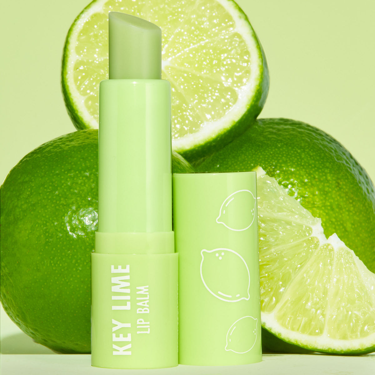 Key Lime ColourPop Lip Balm tube stands uncapped, balm bullet angled up, in glossy light‑green packaging with white “KEY LIME LIP BALM” text and citrus icons, set among whole and sliced limes on a pastel green backdrop.