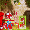 Berry Grinchy So Juicy Plumping Gloss Stick by ColourPop stands upright in a pink tube beside a matching liner and Grinch-themed window box, set among wrapped holiday gifts and ornaments in a cozy festive living room.