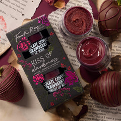 Fourth Ray Beauty Kiss of Darkness lip care kit shows chocolate-covered-strawberry lip scrub and overnight lip mask, jars open to reveal shimmery berry textures, beside the floral black box, styled with chocolate-dipped strawberries on torn book pages; ColourPop Lip Care Kit.