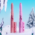 Two Colourpop x Grinch Liked Christmas a Lot… Shadow Stix set - shimmering peach and pink cream eyeshadow sticks with star-print caps - standing in faux snow amid winter trees against a bright blue sky.