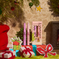 ColourPop Liked Christmas a Lot… Shadow Stix Set displayed upright - two lilac eyeshadow sticks beside a boxed trio in pastel packaging - surrounded by ornaments, wrapped gifts, and a fireplace in a cheerful Grinch-themed holiday scene.