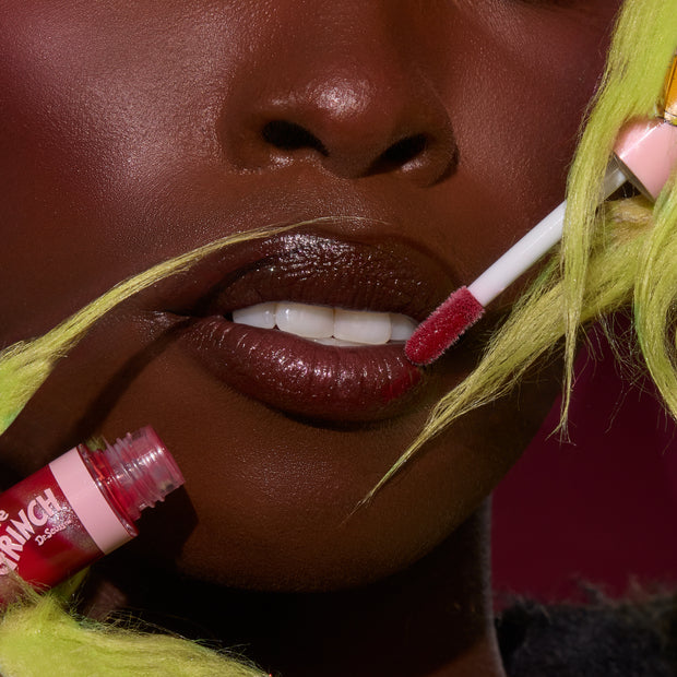 Glossy lips receive a swipe of ruby swirl lip gloss from a doe-foot applicator, with the open red tube nearby, highlighting the ColourPop Merry Grinchmas Swirl Lip Gloss Kit amid lime-green faux fur accents and a deep magenta backdrop.