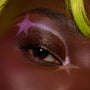 Shimmery bronze and gold flecks glow on a partially open eyelid while a pink‑lilac star graphic wraps the eye, created with the ColourPop Liked Christmas a Lot… Shadow Stix Set; extreme close-up with a neon green strand by the brow.