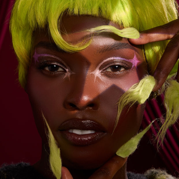 Model wearing ColourPop X The Grinch full collection set, showcasing shimmering eye shadow, graphic white liner, and glossy lips framed by neon green faux fur details against a deep red backdrop, promoting Best of Sitewide ColourPop holiday makeup.