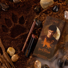 ColourPop Power Pout Lipstick, Bella! Where the Hell Have You Been, Loca?, resting with a matching brown lip pencil and themed box on earthy soil amid stones and driftwood, featuring a wolf paw print and moonlit wolf graphic evoking Twilight New Moon.