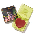 Fairytale Things ColourPop Lip and Cheek Balm sits open in a translucent neon-yellow square compact with an embossed S, revealing a red heart-shaped pan; beside it, the Shrek collab carton shows fairytale characters on a white studio background.