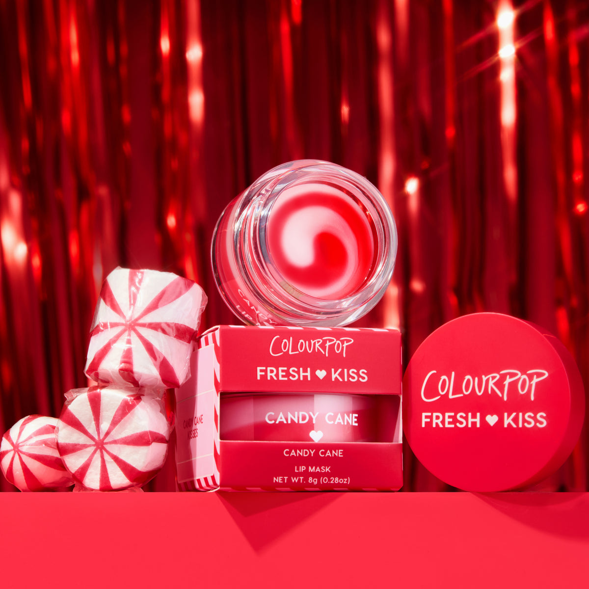 ColourPop Fresh Kiss Candy Cane Lip Mask jar opens to show glossy red balm, stacked with its red 0.28 oz boxes beside peppermint candies, set against a shiny red tinsel holiday backdrop for a festive, gift-ready beauty shot.