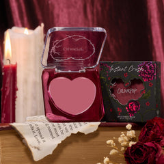 Heart-shaped Love Interest compact open to reveal rosy mauve balm, displayed with floral “Instant Crush” box, lit candles, a torn book page, and dried roses on a wood table - ColourPop Lip and Cheek Balm for lips and cheeks.