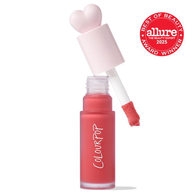 ColourPop Liquid Blush She’s Cute shown open, doe-foot applicator lifted from a frosted coral-pink bottle with a pale pink heart-shaped cap, revealing creamy formula on the tip; white backdrop with an Allure 2025 Best of Beauty award badge.