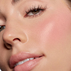 ColourPop Lip and Cheek Balm in Lucky U delivering a dewy soft-pink flush on the cheek and a glossy nude tint on the lips, shown in an extreme close-up of a face with smooth skin and long lashes under natural light.