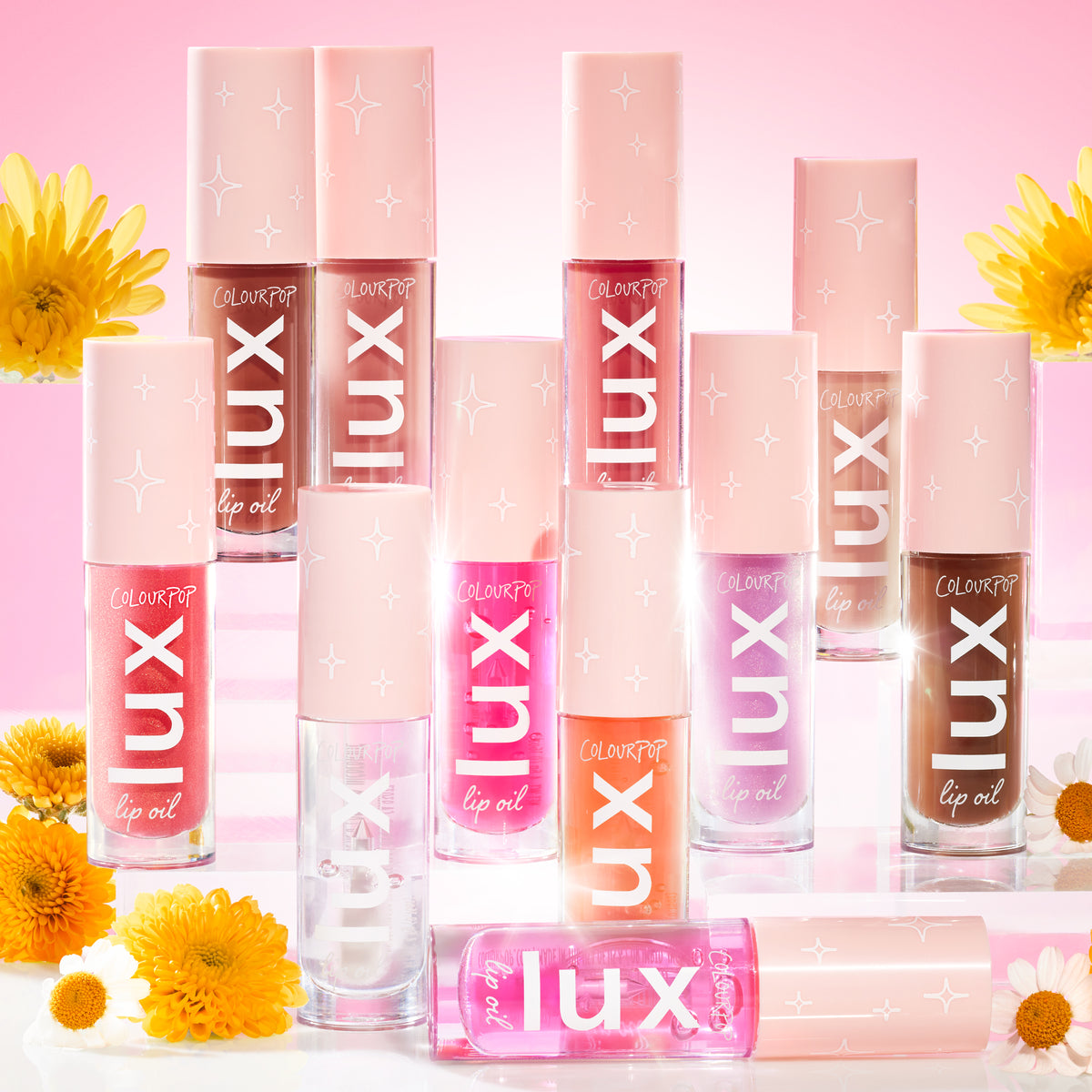 ColourPop Lux Lip Oil bottles from the Comb By collection stand in rows, peach caps with white star icons and bold LUX lettering, against a rosy backdrop with daisies, showing clear, coral, pink, mauve, and deep brown shades for glossy, hydrating lips.