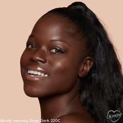 Smiling model showcases ColourPop Pretty Fresh Creamy Concealer in Deep Dark 220 C, revealing smooth, radiant, skin-like coverage; close-up studio portrait with glossy lips, high ponytail, and peach background with ColourPop logo.