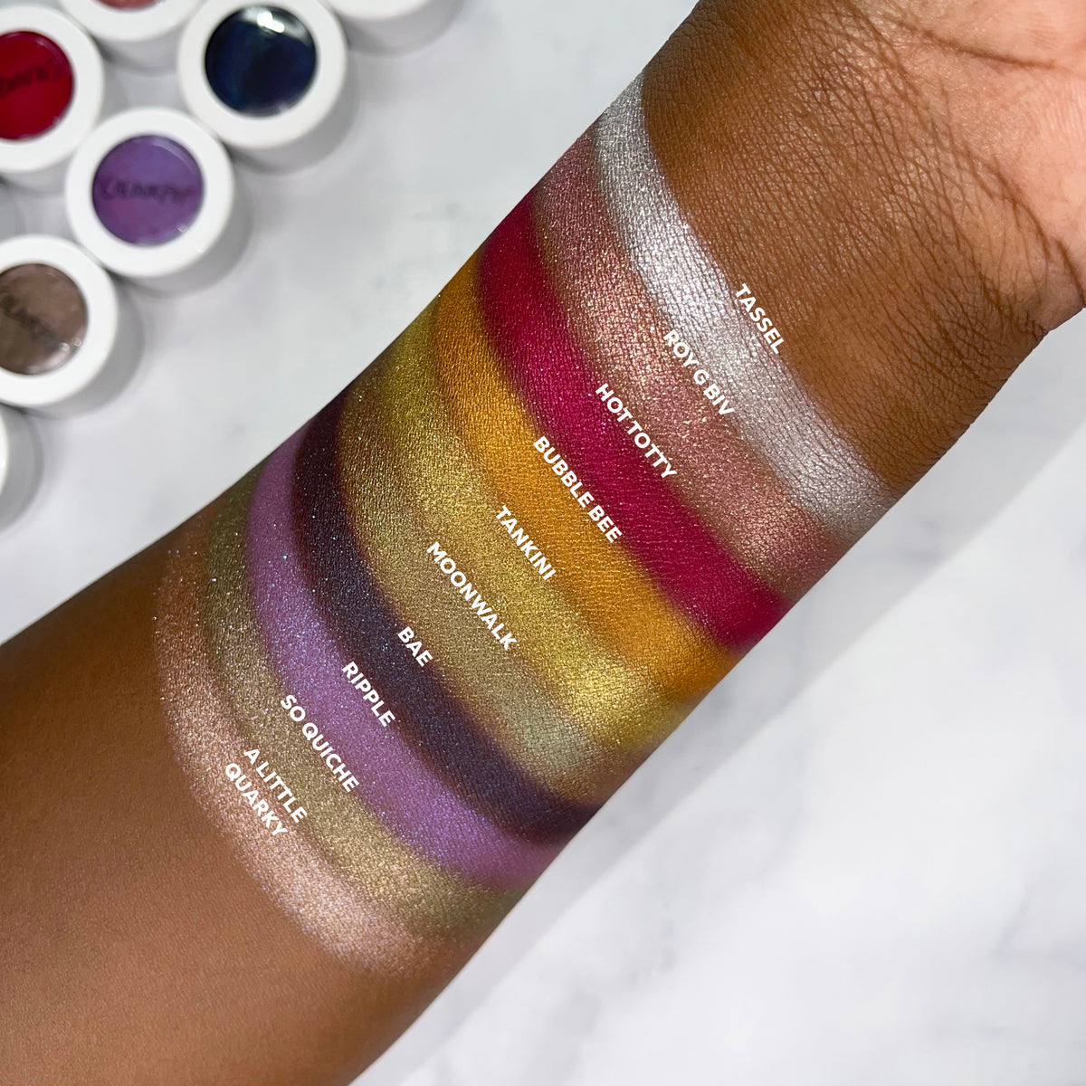 Bae Super Shock Shadow by ColourPop swatched on an arm, showing a deep plum base with vivid blue shimmer, displayed among labeled Super Shock Shadow swatches with ColourPop pots blurred in the background.
