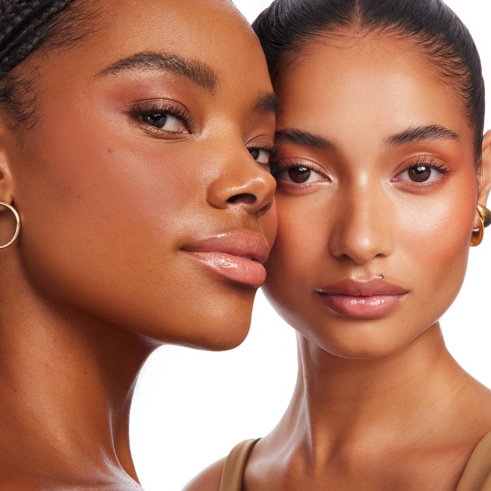 Two models showcase a sun-kissed glow, wearing La Jolla Cove ColourPop Bronzer Stix blended on cheeks and temples, with dewy skin, glossy lips, and small gold hoops in a close-up studio shot against a white background.