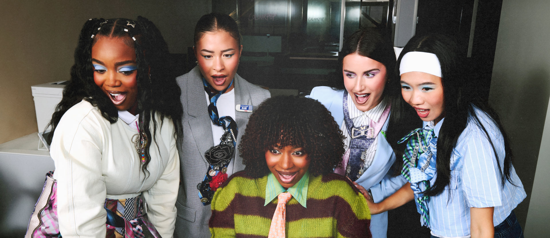 Group of five stylish young women with bold, colorful ColourPop-inspired office makeup crowd around a computer, reacting excitedly in a modern workplace setting.