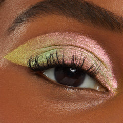 Close-up eye wearing Galaxy Shadow Stix eyeshadow, showing a bold cut-crease look in metallic pink, green, and gold shimmer on deep skin, highlighting the multi-chrome pigment and smooth, blendable finish for dramatic glam makeup.